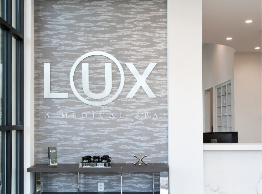 Reception area of LUX Medical Spa, showcasing a modern gray feature wall with large illuminated “LUX A MEDICAL SPA” signage and a sleek console table with branded products at the entrance.