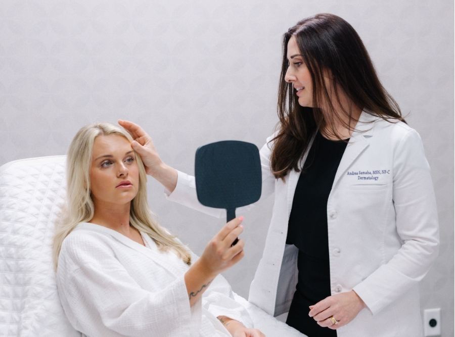Dermatology provider in a white coat offering medical oversight to a spa patient, as the patient sits on a treatment bed in a robe and evaluates her results in a handheld mirror.