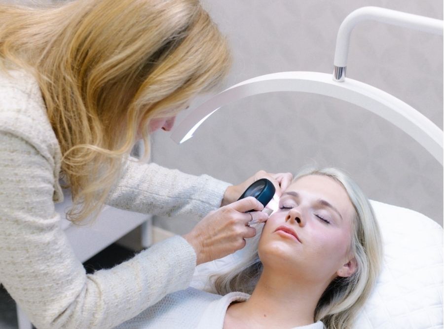 Skin care specialist performing a personalized facial consultation on a patient at lux medical spa, examining the client’s skin under a bright treatment light while the patient reclines comfortably.
